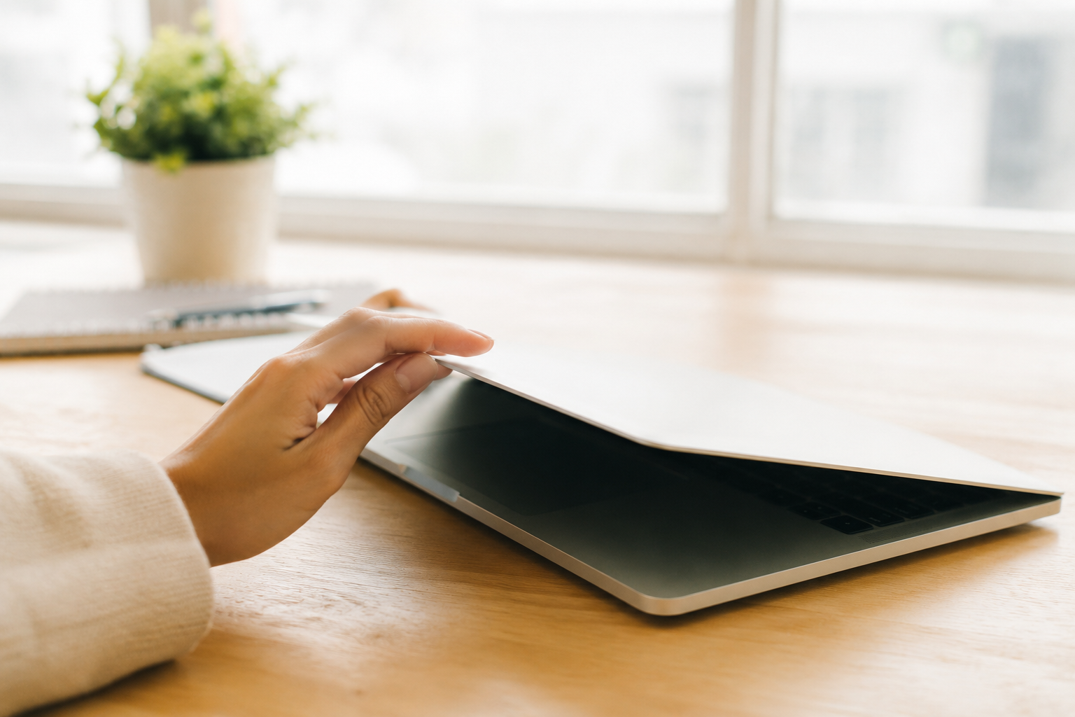 Hand closing a laptop on a wooden desk beside a notebook and plant, representing the start of a digital detox